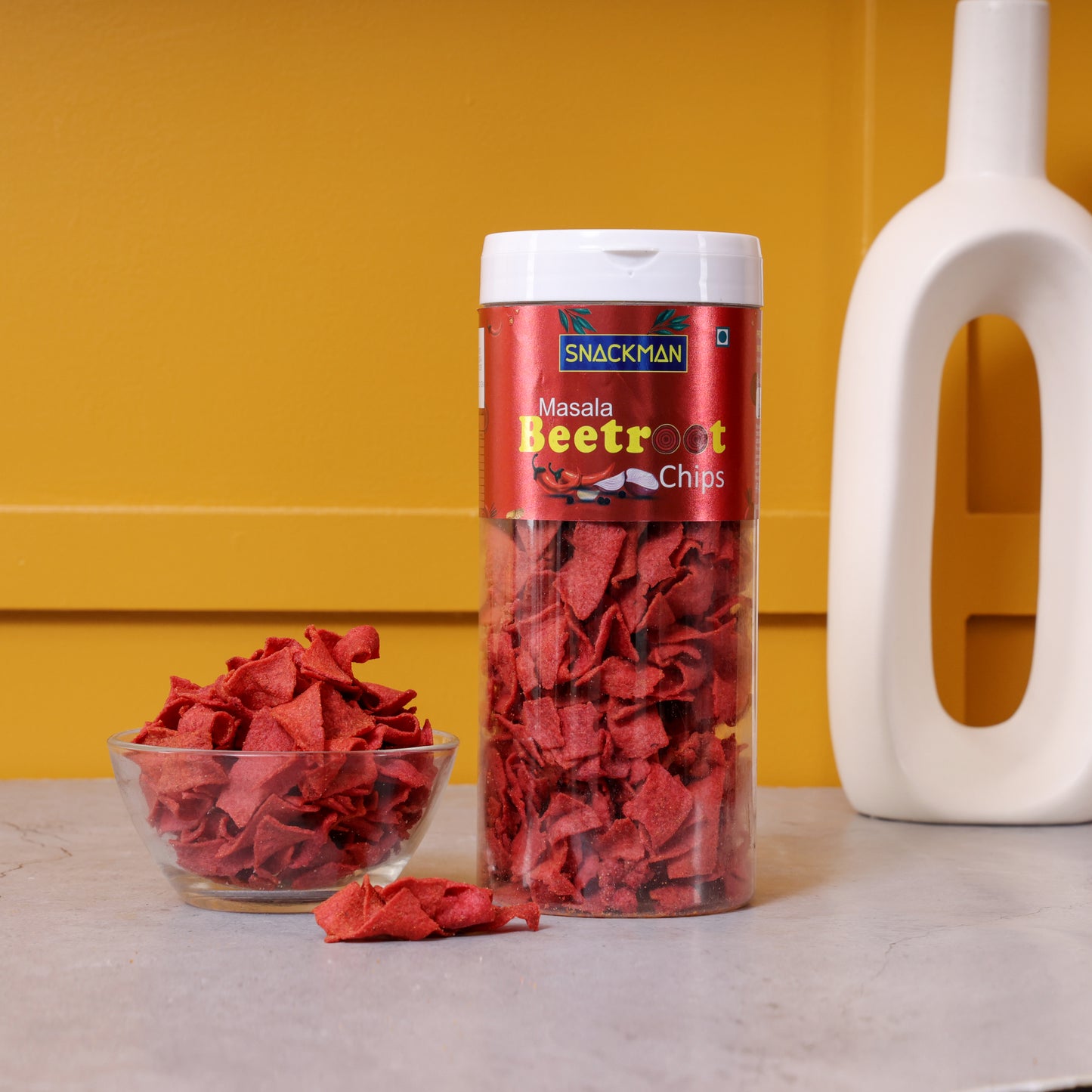 snackman masala beetroot chips in a cylindrical container with a bowl of chips on a yellow background
